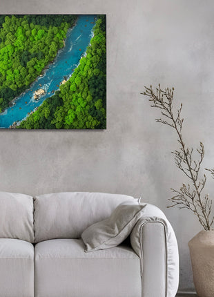 Framed artwork of a river surrounded by greenery above a gray sofa with a plant on a matching side table.