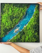 Framed artwork of a river surrounded by green trees held by hands on a white background