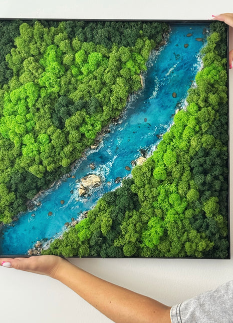 Framed artwork of a river surrounded by green trees held by hands on a white background