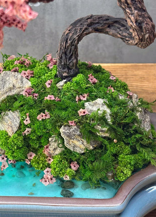 Bonsai-style arrangement with greenery, rocks, and pink flowers on a blue tray.